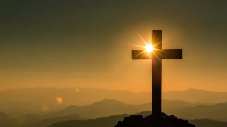 Background mountains with sun coming up behind a cross in the foreground.